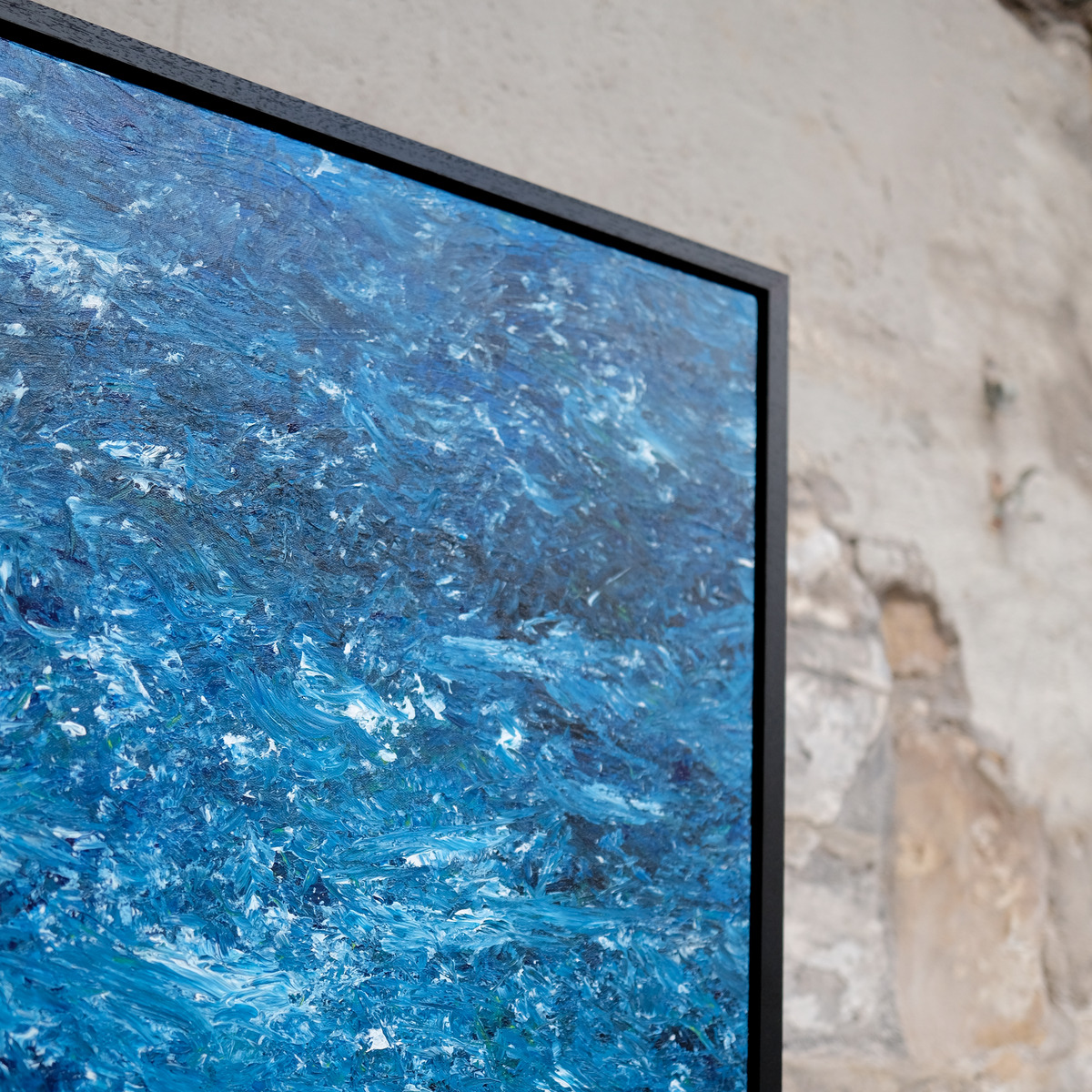 Angled close-up of the upper-right corner of A Thousand Tides, showing densely textured blue paint, scattered white marks, canvas texture and the black frame.